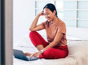 A young woman sitting on a bed is resting her arm on her knee an A young woman sitting on a bed is resting her arm on her knee and smiling down are her open laptop.