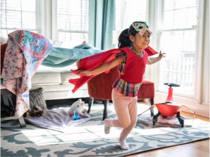 A young girl wearing a cape on her back and a butterfly mask on A young girl wearing a cape on her back and a butterfly mask on her head is smiling while running forward with her arms stretched out.