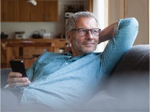 A middle-aged man holding a smartphone is wearing glasses while A middle-aged man holding a smartphone is wearing glasses while sitting on a couch with his arm tucked behind his head while he looks out to his left.